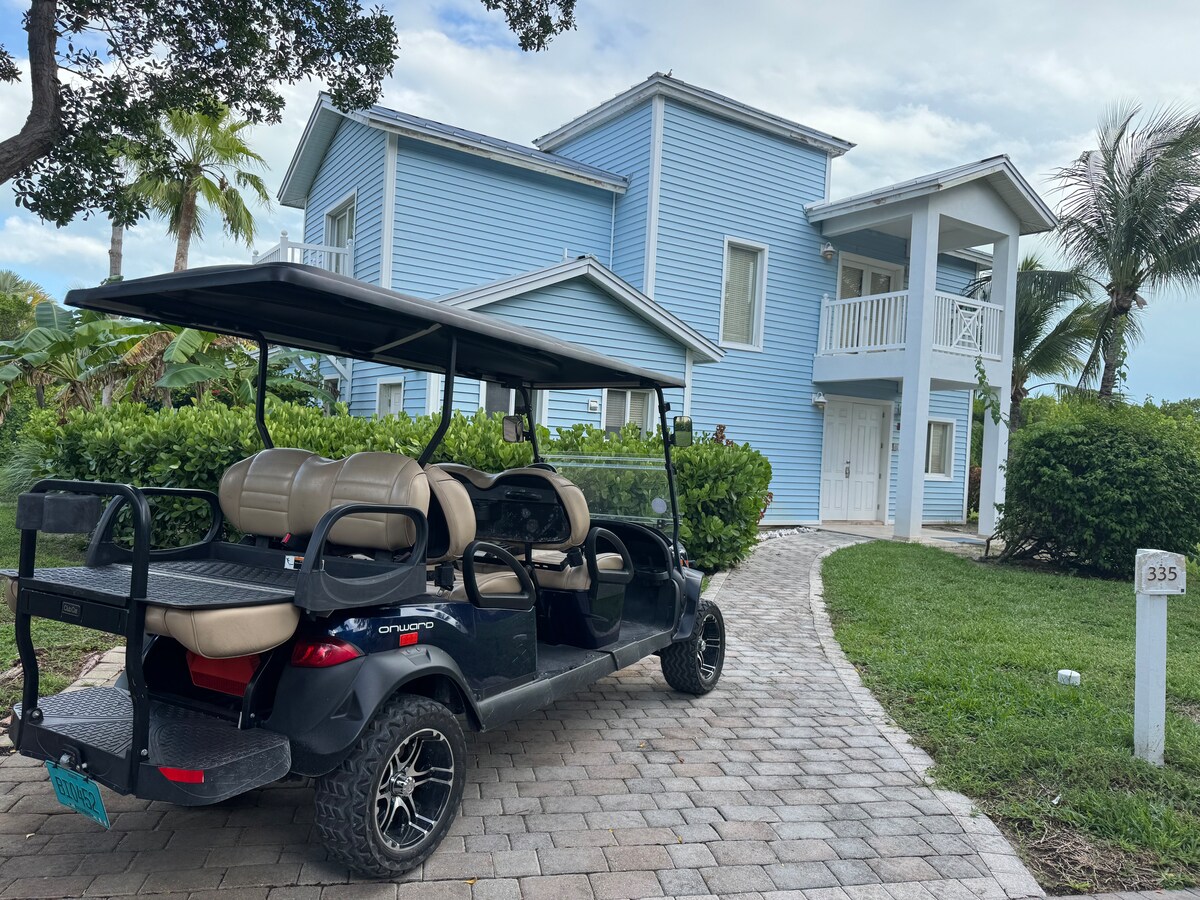 A golf cart is parked along a stone pathway leading to a light blue, two-story house surrounded by greenery. Multiple palm trees and shrubs are seen enhancing the tropical setting. The home features a prominent balcony on the upper floor, offering a view of the surroundings.