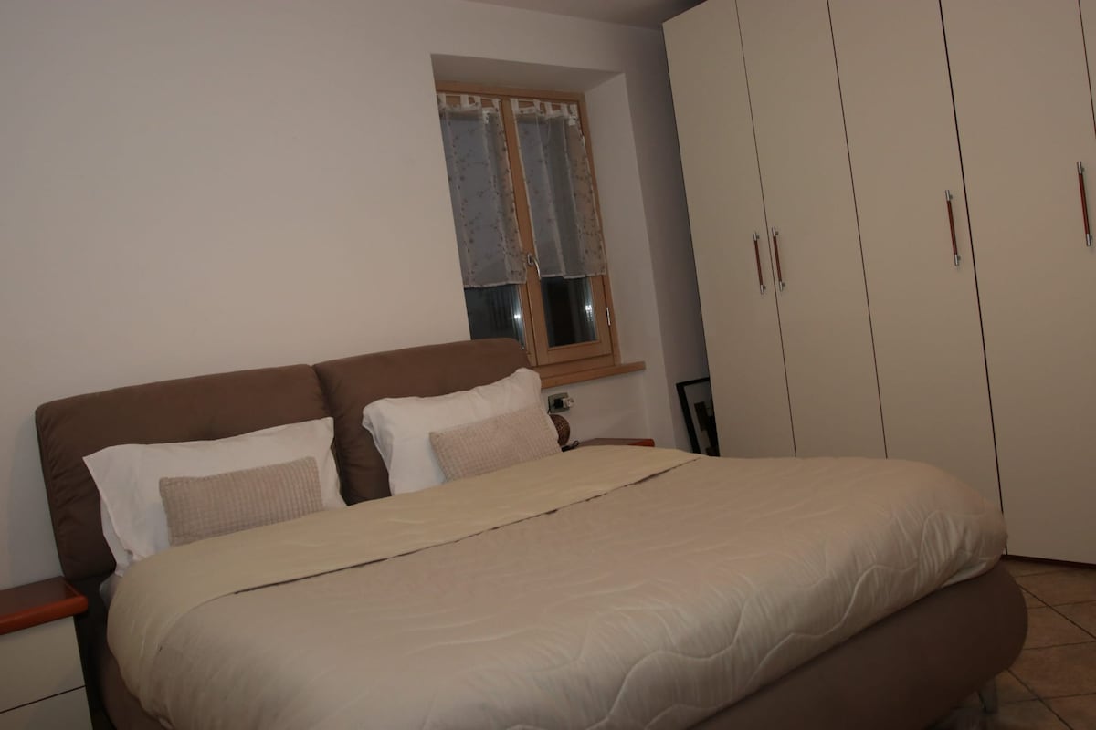 A spacious bedroom is presented, featuring a large bed with neutral bedding and two pillows. A pair of bedside tables are placed on either side of the bed. Natural light enters through a window adorned with lace curtains, while a built-in wardrobe occupies one wall.