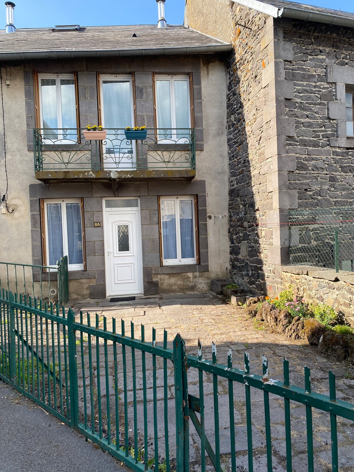 The front exterior of a two-story stone house is visible, featuring a white door framed by two windows. A narrow balcony adorned with flower boxes is located on the upper floor. A green gate and a pathway lead to the entrance, surrounded by a small garden area.