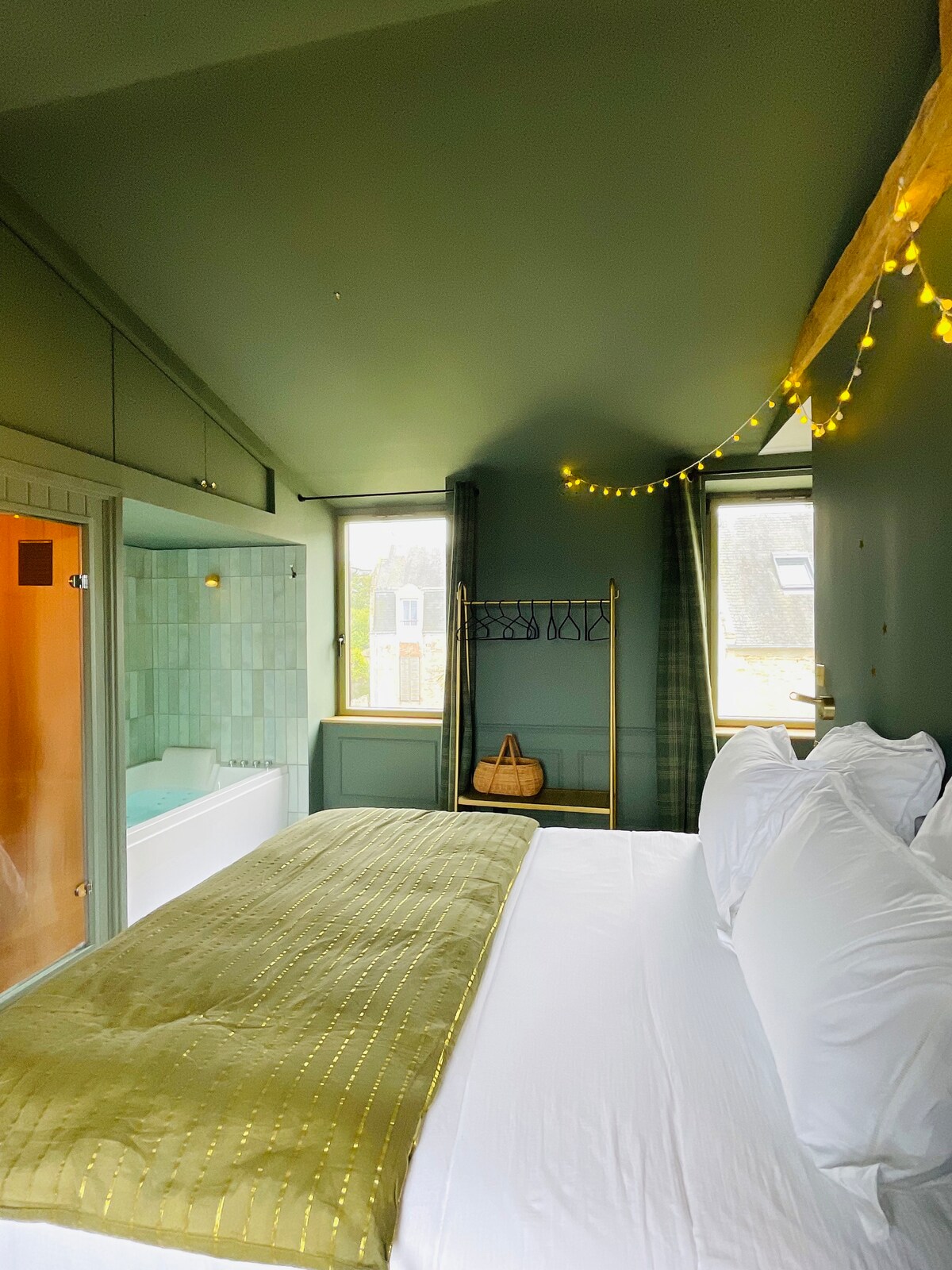 A spacious bed sits adorned with a light green throw, framed by large windows allowing natural light to fill the room. Subtle fairy lights provide a warm glow, and a glimpse of an elegant bathroom is visible in the corner.