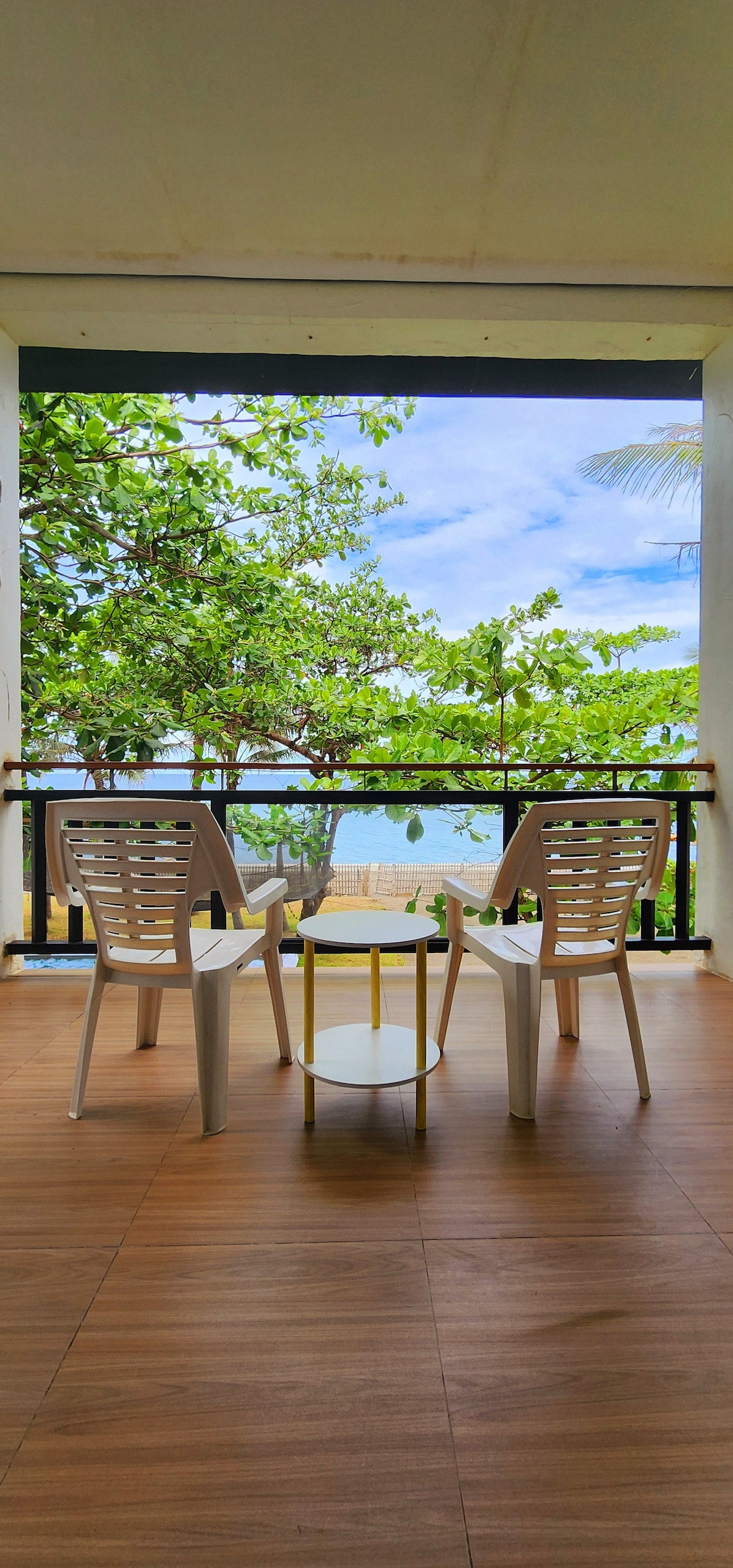 A serene outdoor seating area features two white chairs positioned before a small round table. Lush greenery frames the view, with glimpses of the beach and ocean visible in the background, inviting relaxation and enjoyment of the coastal surroundings.