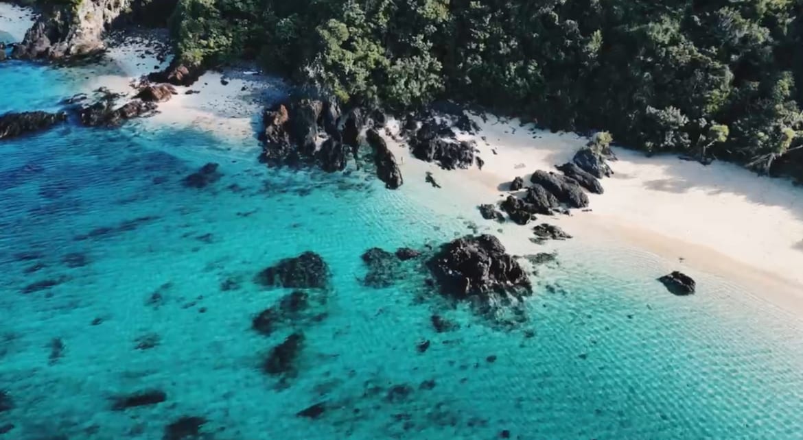 A serene beach scene is presented, showcasing soft white sand bordered by clear turquoise waters. Rocky formations are visible along the shoreline, with lush greenery providing a natural backdrop. The gentle waves create a tranquil atmosphere, inviting exploration of the coastline.
