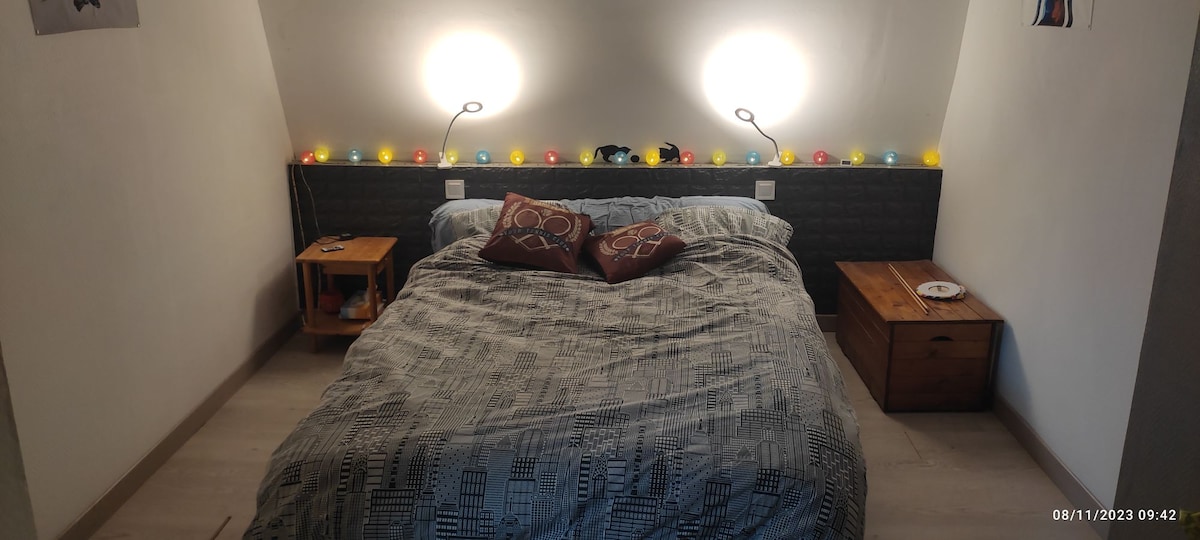 A cozy bedroom features a large bed adorned with patterned bedding. Two modern bedside tables are present, each holding a small item. Soft, illuminated decorative lights line the wall behind the headboard, providing a warm ambiance.