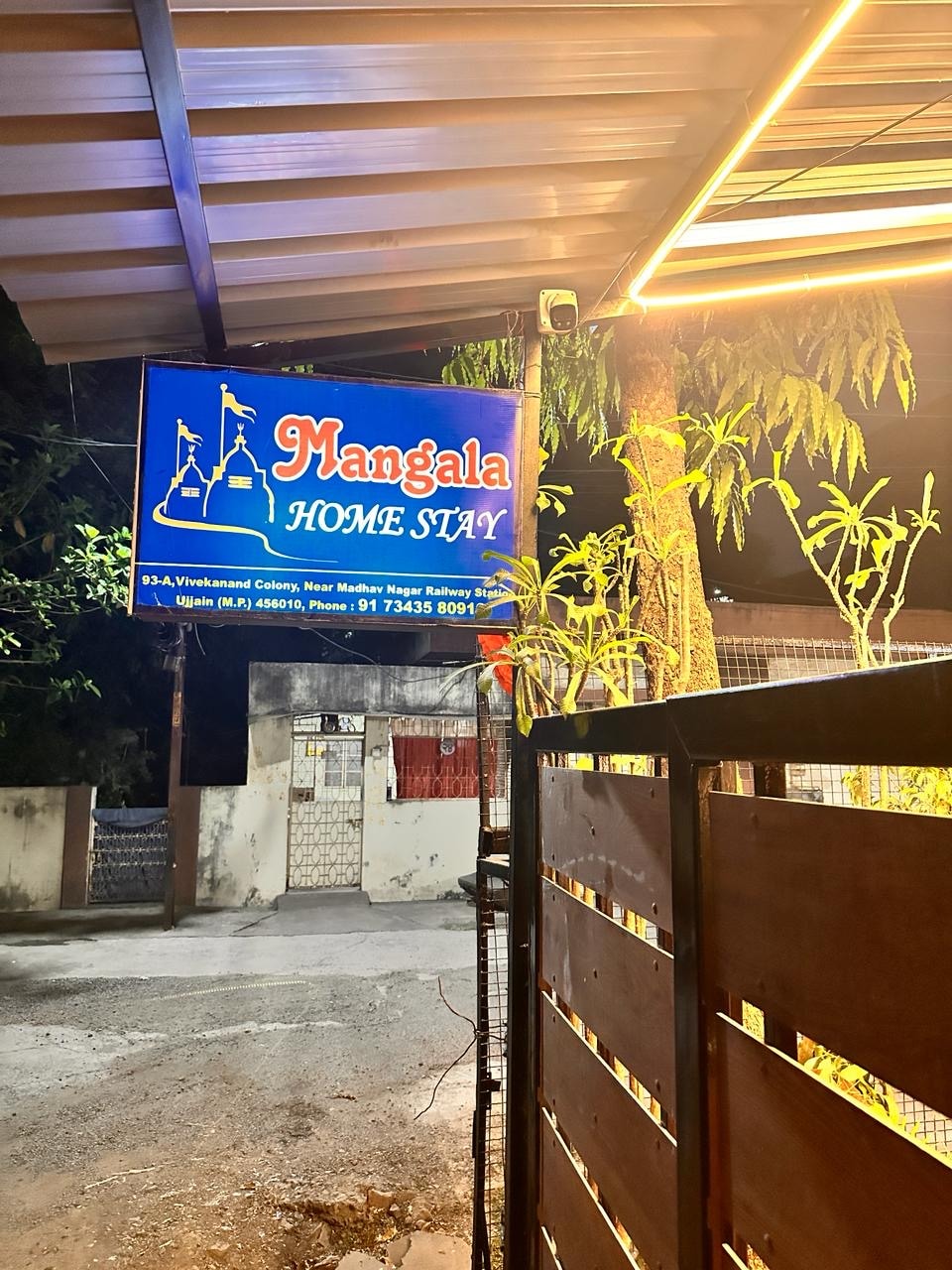 A illuminated sign for Mangala Home Stay is displayed prominently, featuring a nautical theme with a ship graphic. The surrounding foliage enhances the welcoming entry, with a wooden fence providing a boundary. The scene is set during nighttime, with soft light accentuating the area.