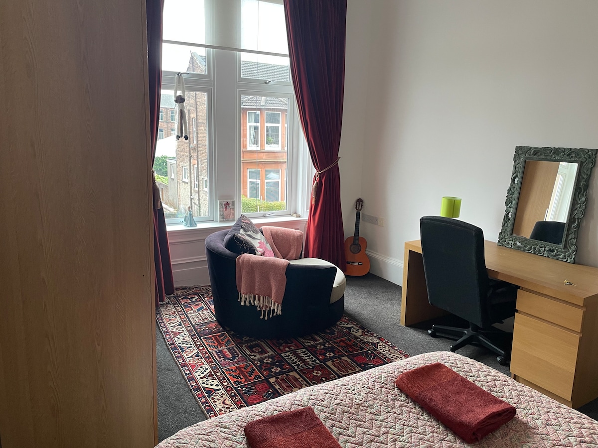 A well-lit room features a large window with red drapes, allowing natural light to fill the space. A comfortable chair with a soft throw is positioned near the window, alongside a desk and mirror. A patterned rug adds warmth, while a glimpse of the outside can be seen.