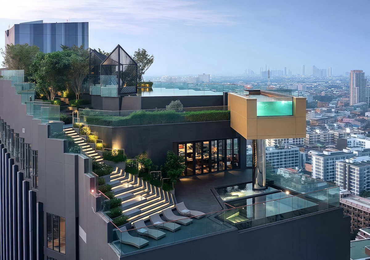 A rooftop area features a spacious infinity pool overlooking the city. Lounge chairs are arranged along the terrace, while green trees and plants are strategically placed, adding touches of nature. Modern architecture is showcased in the adjoining lounge area with large glass windows.