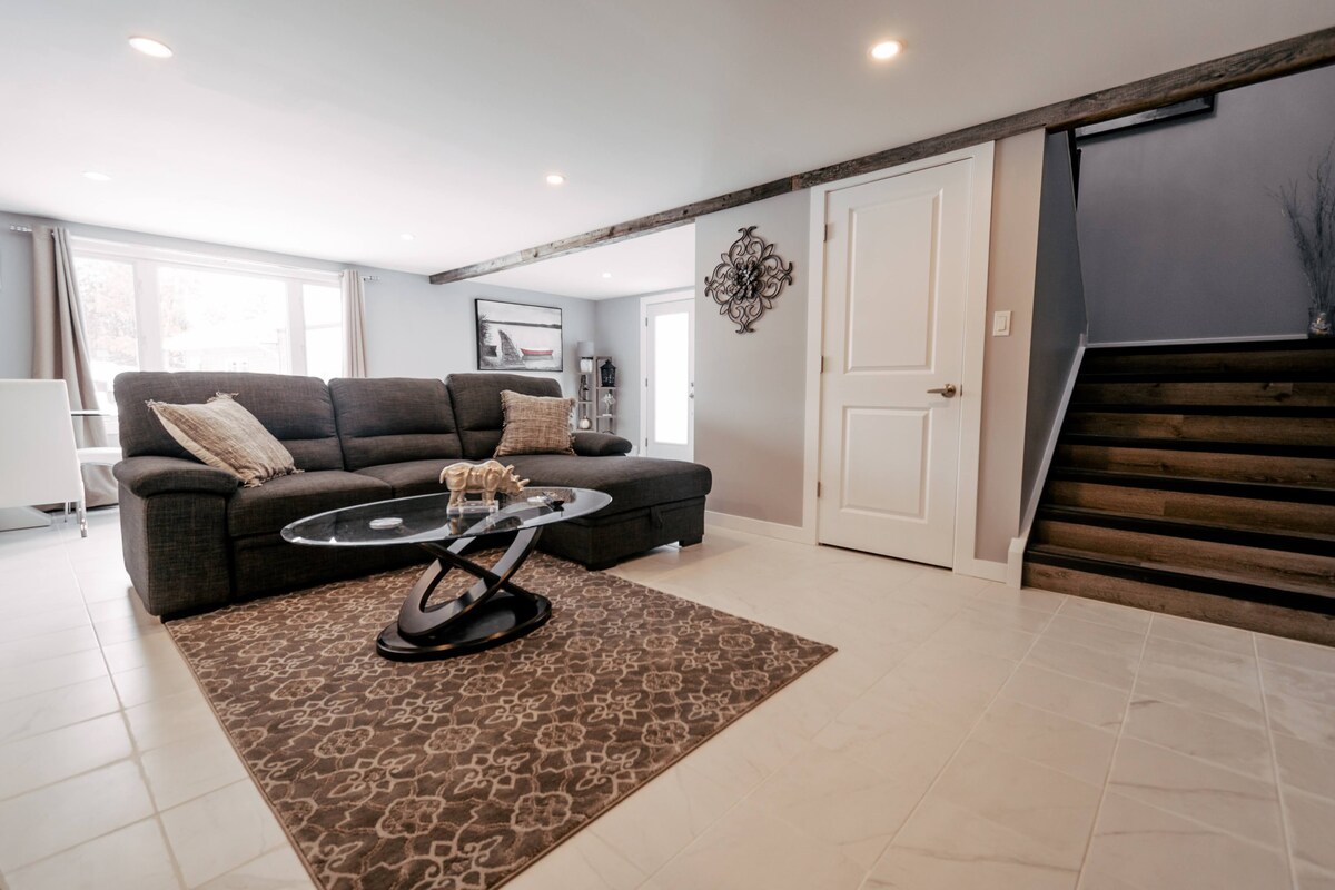 A modern living area is showcased, featuring a spacious sectional sofa paired with a sleek glass coffee table. A decorative area rug adds warmth to the tiled floor. Natural light fills the room through large windows, and a staircase leads to the upper level.