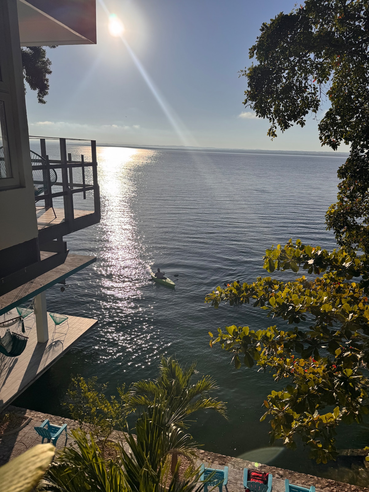 A tranquil view over Lake Petén Itzá is captured, with sunlight reflecting off the water's surface. Green foliage frames the scene, and a pathway leads to a deck adorned with coordination of outdoor seating, inviting relaxation by the serene lakeside.