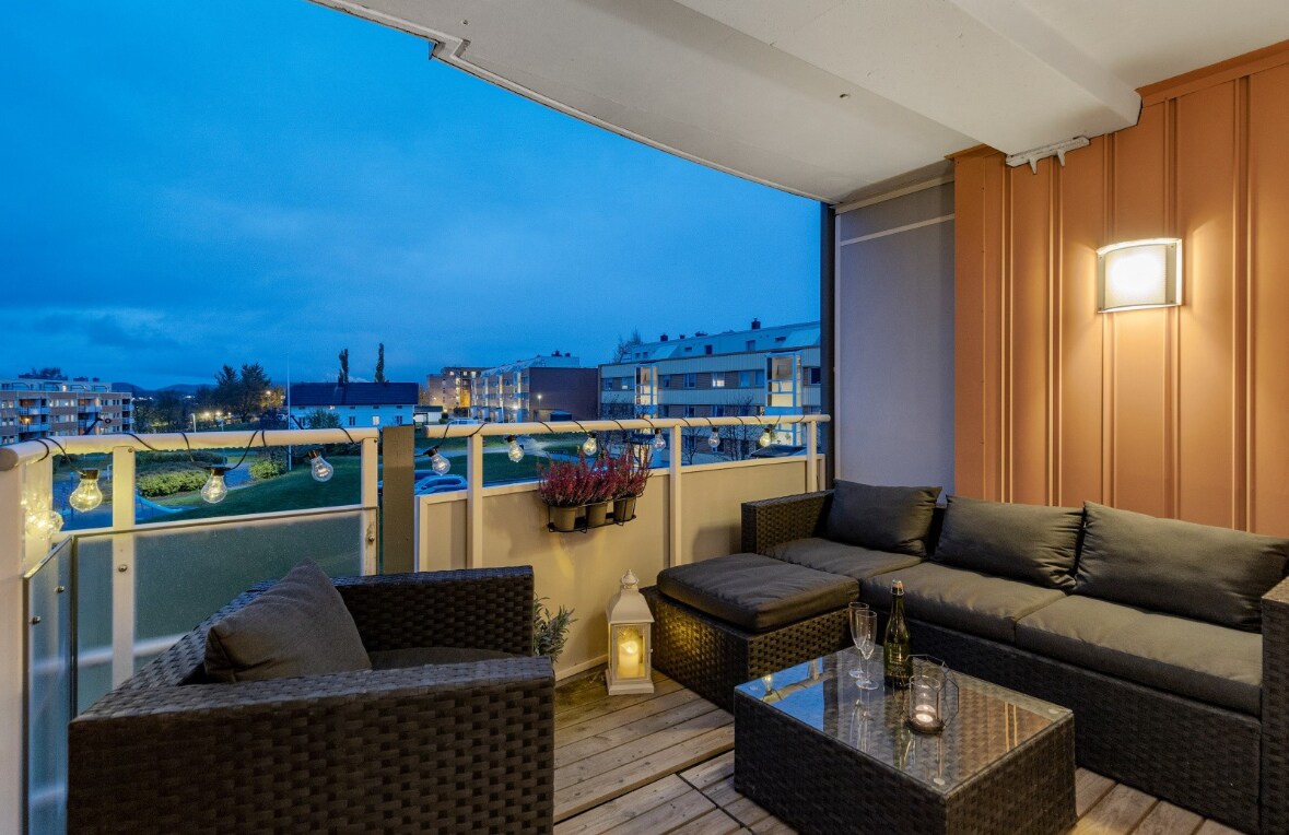 An outdoor terrace features comfortable seating arrangements with dark wicker furniture. A glass table sits in the center, complemented by decorative lanterns. Subtle lighting enhances the relaxing atmosphere, while city buildings are visible in the background under the evening sky.
