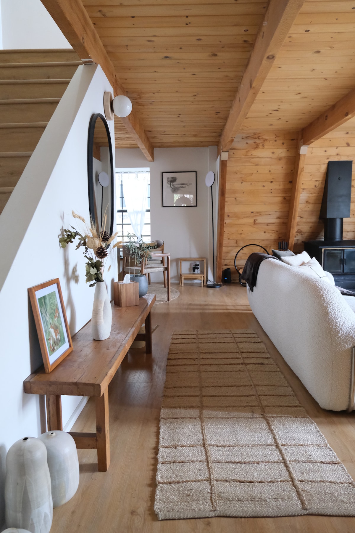 The living room features a textured area rug over wooden flooring, a comfortable couch, and natural wooden accents. A mirrored wall hangs above a simple wooden bench, while a cozy chair and framed artwork complete the inviting space, illuminated by natural light from a large window.