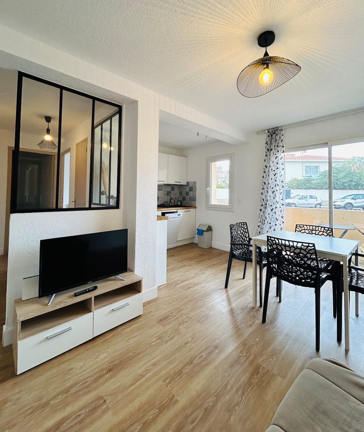 A modern living area features a television on a light wood cabinet, with a dining table surrounded by four chairs. Bright light enters through large windows, highlighting the open layout and warm wood flooring. A view of the outside is visible through glass doors.