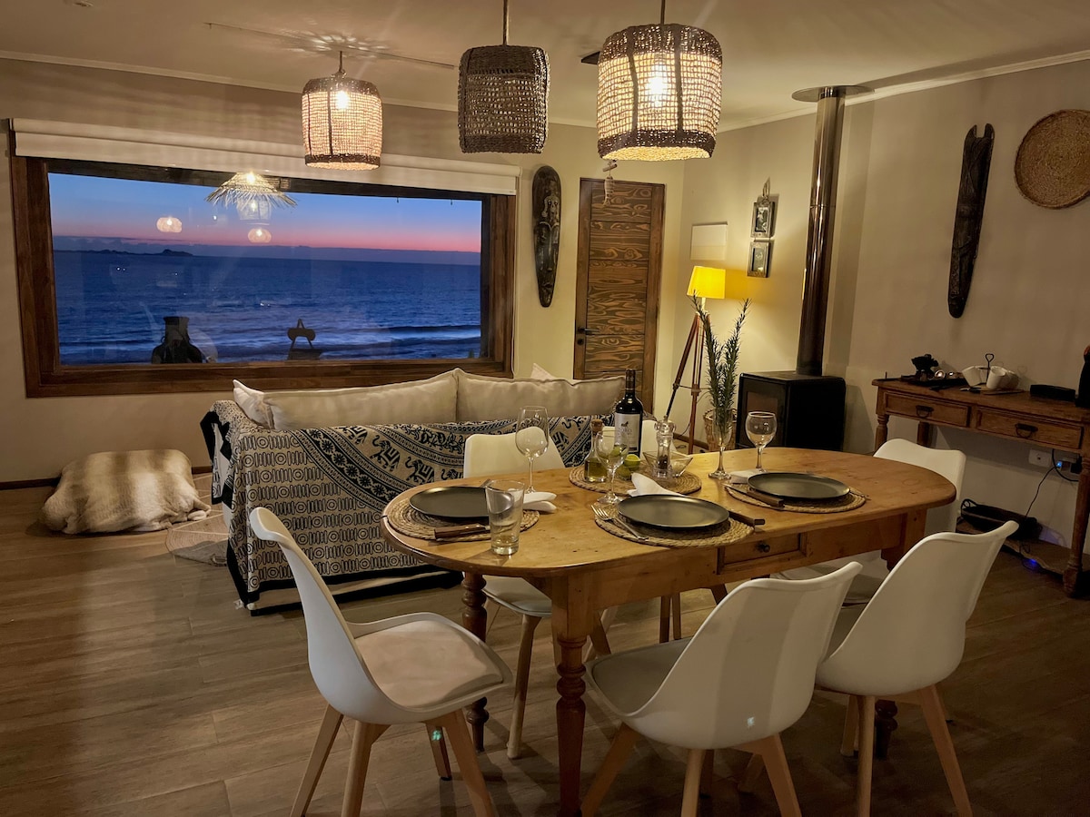 A dining area is highlighted by a wooden table set for a meal. Modern chairs surround the table, while a comfortable seating area with cushions is nearby. Large windows frame a sunset view over the ocean, enhancing the warm atmosphere of the space.