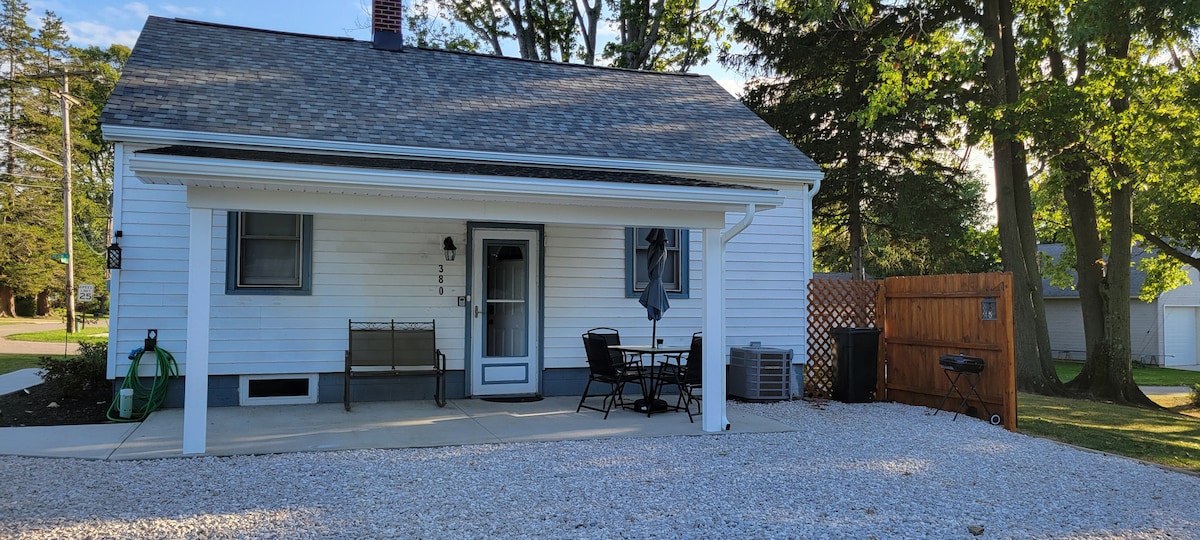 The exterior of a single-story house features a covered front porch with a bench and a small dining table. A gravel walkway leads to the entrance, surrounded by grassy areas and trees. A grill is positioned near a wooden fence, with ample parking space available.