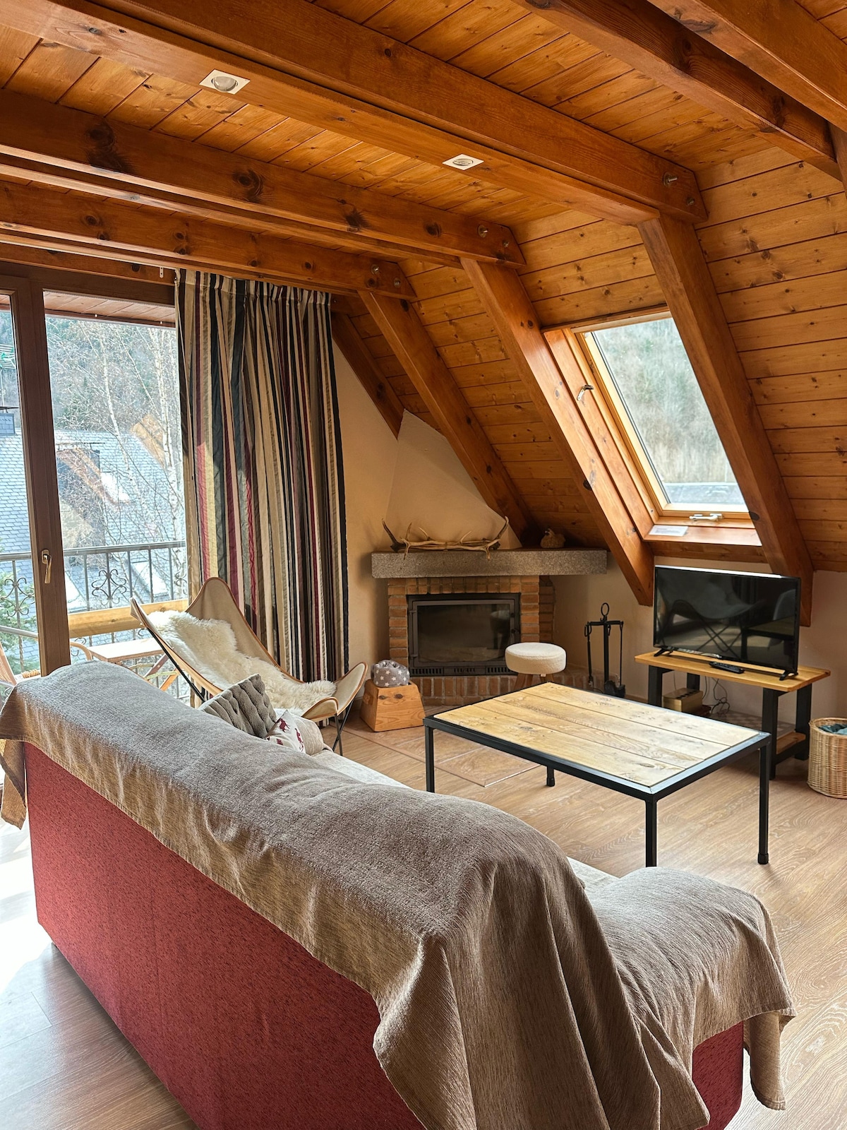 A spacious living area is characterized by wooden beams and sloped ceilings. A comfortable sofa draped with a light blanket faces a wooden coffee table. A small television is positioned next to a decorative fireplace. Large windows provide natural light and views of the surrounding area.