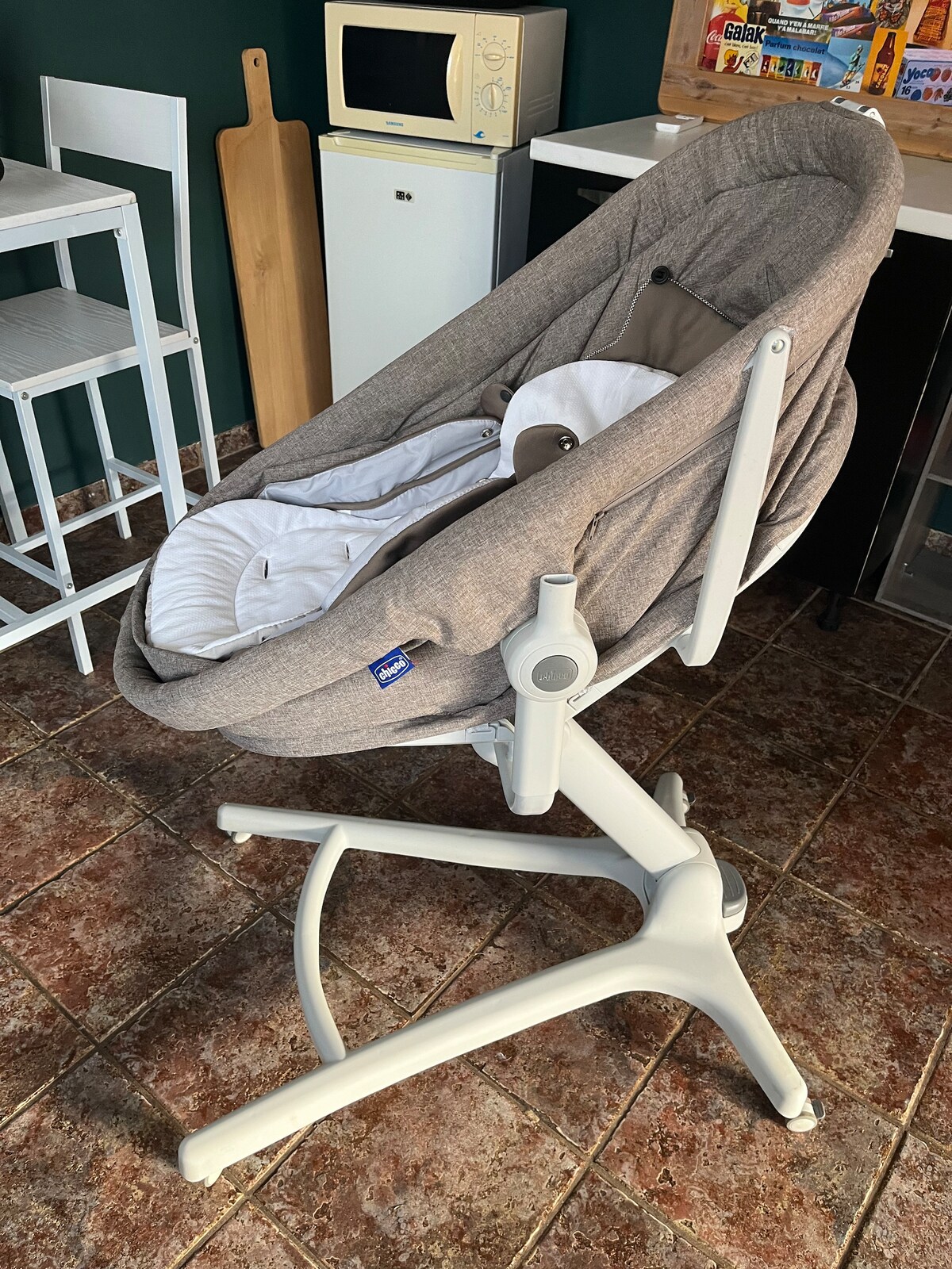 A modern baby bassinet is positioned on a stable white frame. The soft, light grey fabric is complemented by a plush white mattress. Nearby, a small kitchenette with essential appliances, including a microwave, is partially visible. The floor features dark, textured tiles.