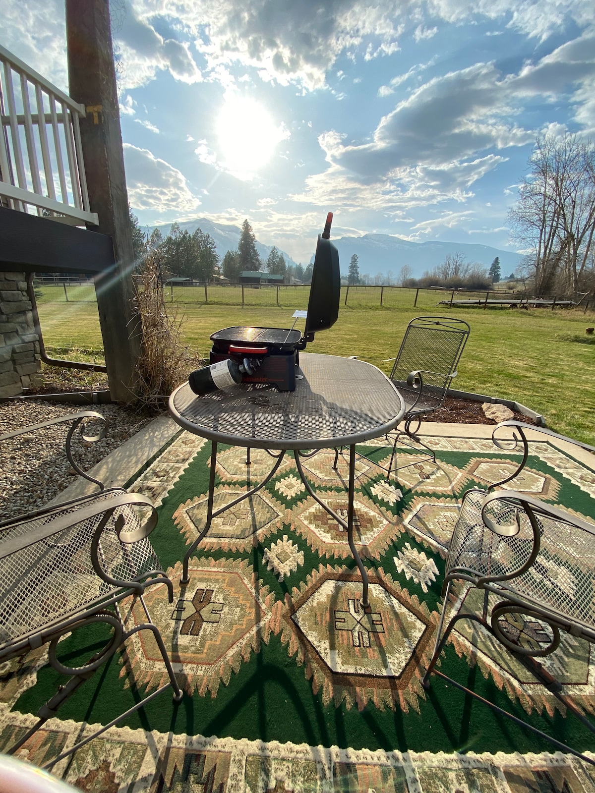 A metal table is positioned on a patterned rug, surrounded by multiple chairs. A portable grill is placed centrally on the table. Natural light illuminates the scene, highlighting the grassy outdoor area and distant trees under a partly cloudy sky.