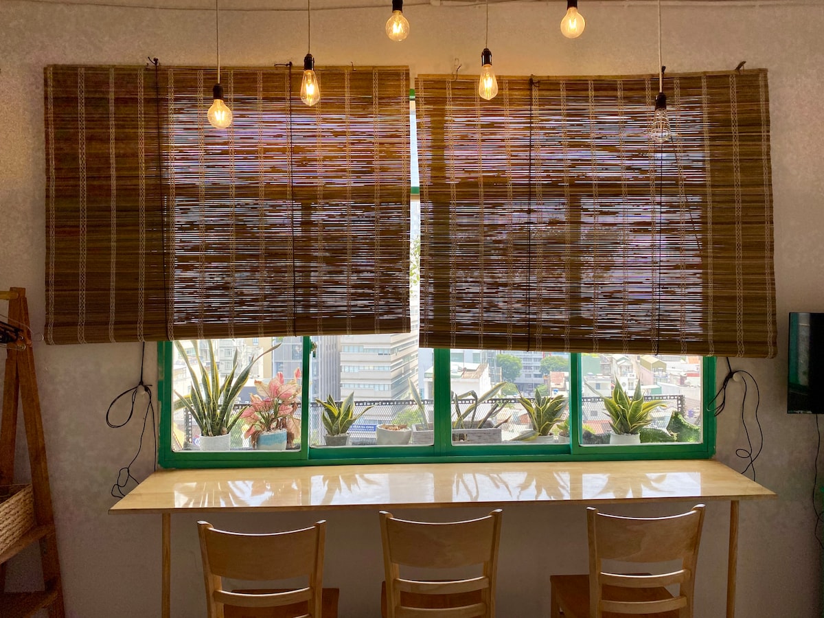 A window area features bamboo blinds allowing natural light to filter through. Below, a light-colored table accommodates several wooden chairs. Potted plants are arranged on the windowsill, adding a touch of greenery to the space, while hanging light bulbs provide illumination.