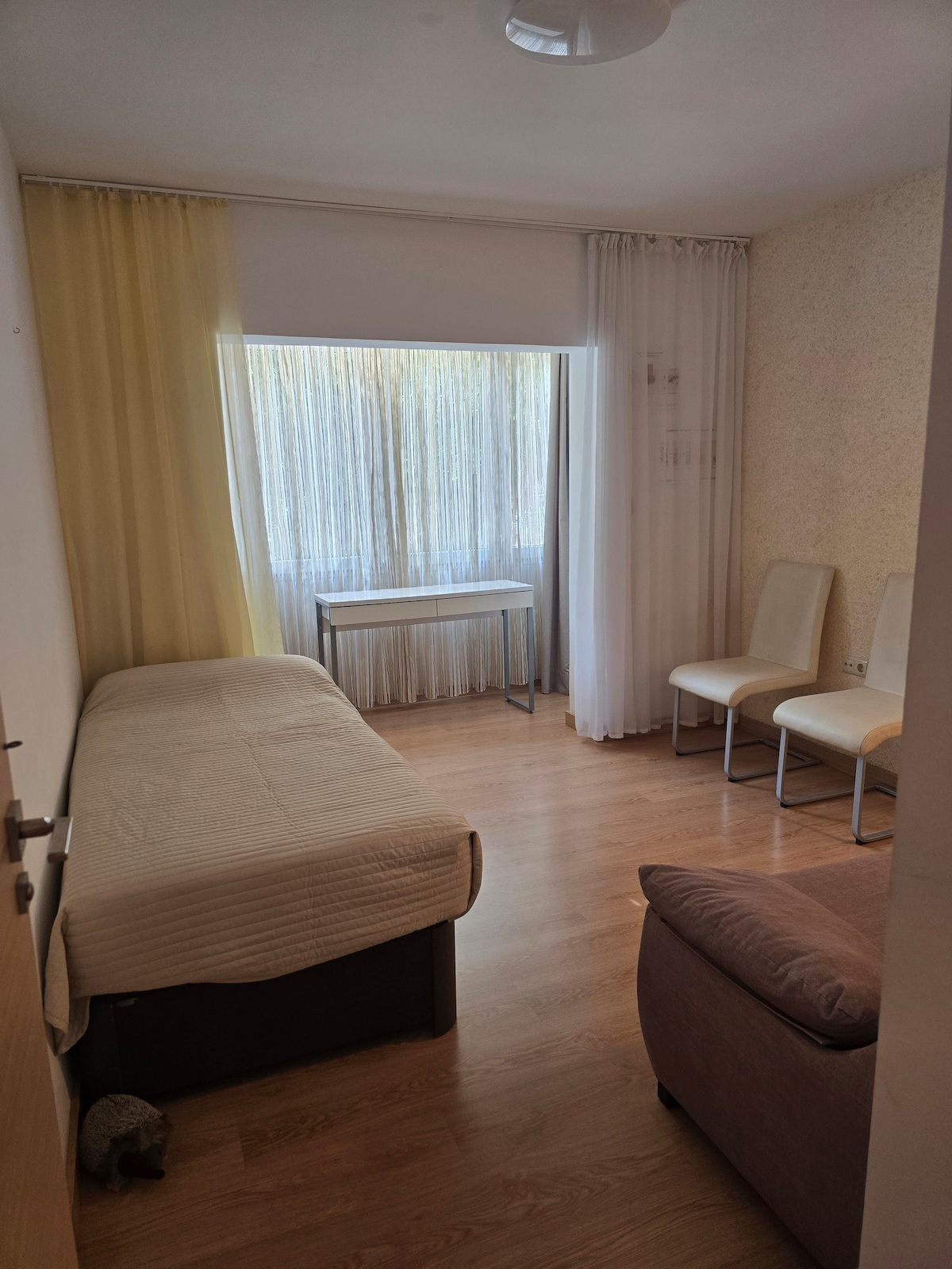 A bright and airy room features a single bed with a neutral-toned coverlet. A small table is situated beside large windows adorned with sheer curtains, allowing natural light to fill the space. Two light-colored chairs accompany a cozy sofa in the seating area.