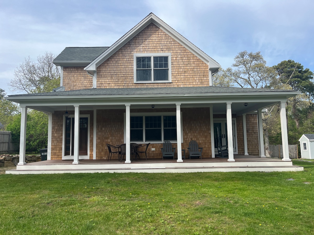 The exterior of a charming two-story house is shown, featuring a wrap-around porch with multiple seating options. The porch is adorned with wooden chairs, and the front yard is grassy, providing ample space for outdoor relaxation. Large windows reflect natural light, contributing to the inviting ambiance.