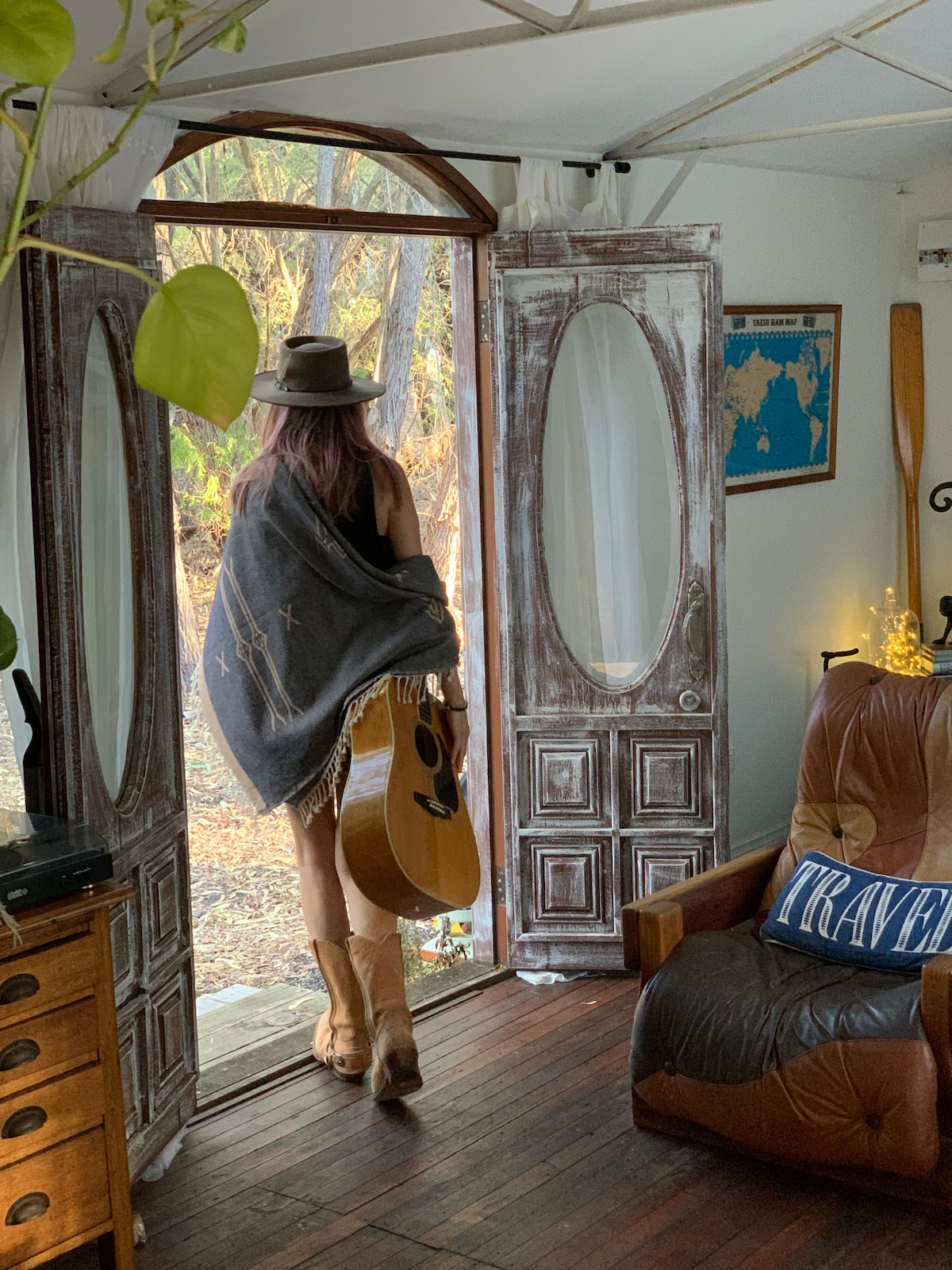 A figure holding a guitar stands in the doorway, wearing a hat and a cozy shawl. The rustic double doors are open, framing a view of the natural surroundings. Light wood floors and a comfortable chair are visible inside, decorated with a travel-themed pillow.
