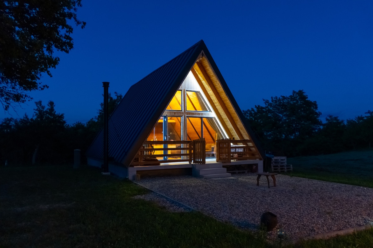 An A-frame house is illuminated under a night sky, showcasing a warm glow from large windows. The exterior features a sloped roof and wooden accents. A spacious outdoor area includes gravel pathways and seating, surrounded by greenery and trees.