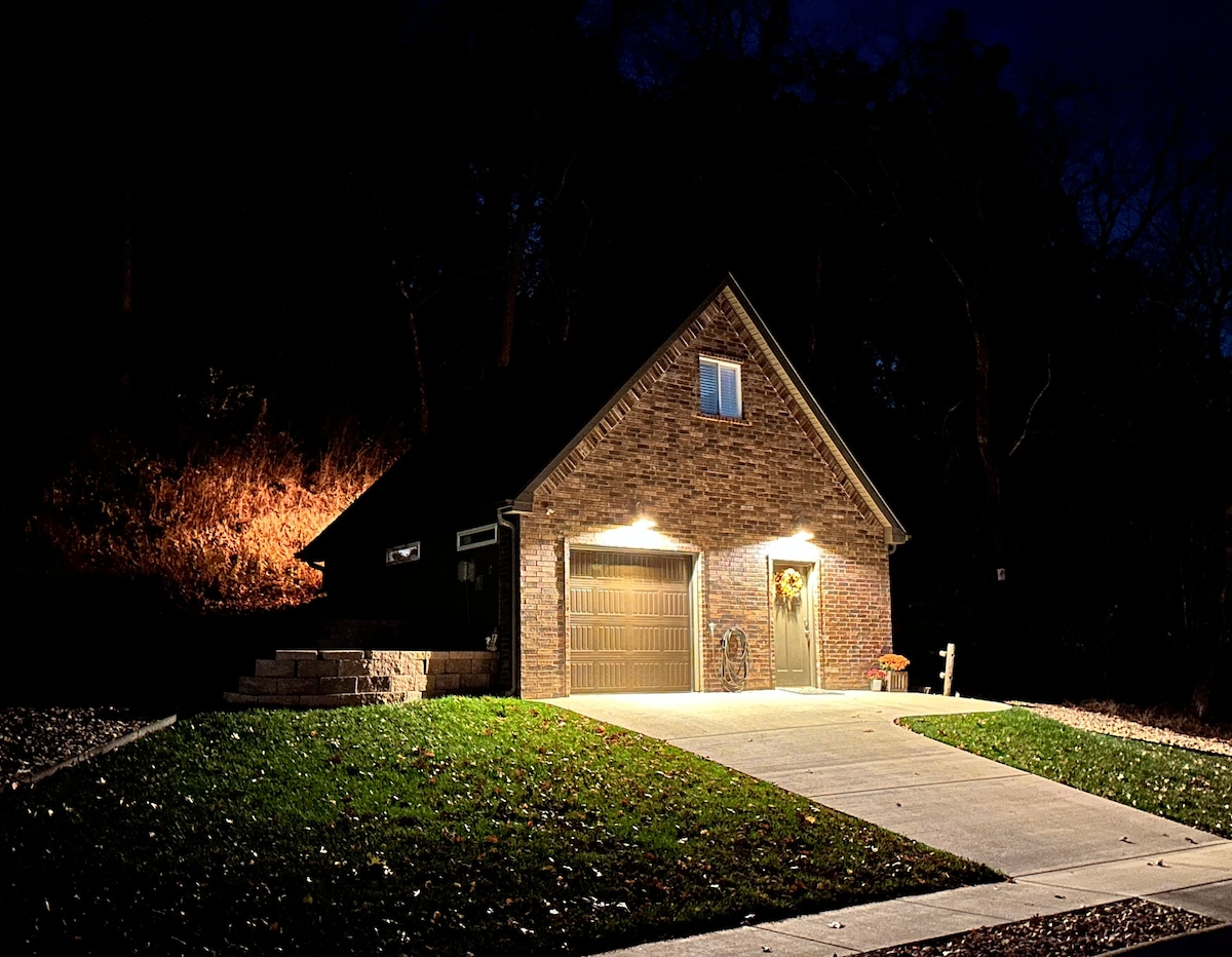 The exterior of a brick structure is illuminated by warm lights, creating a welcoming presence at night. A driveway leads up to the building, which features a small garage and a decorated front door. Surrounding greenery adds to the serene setting.