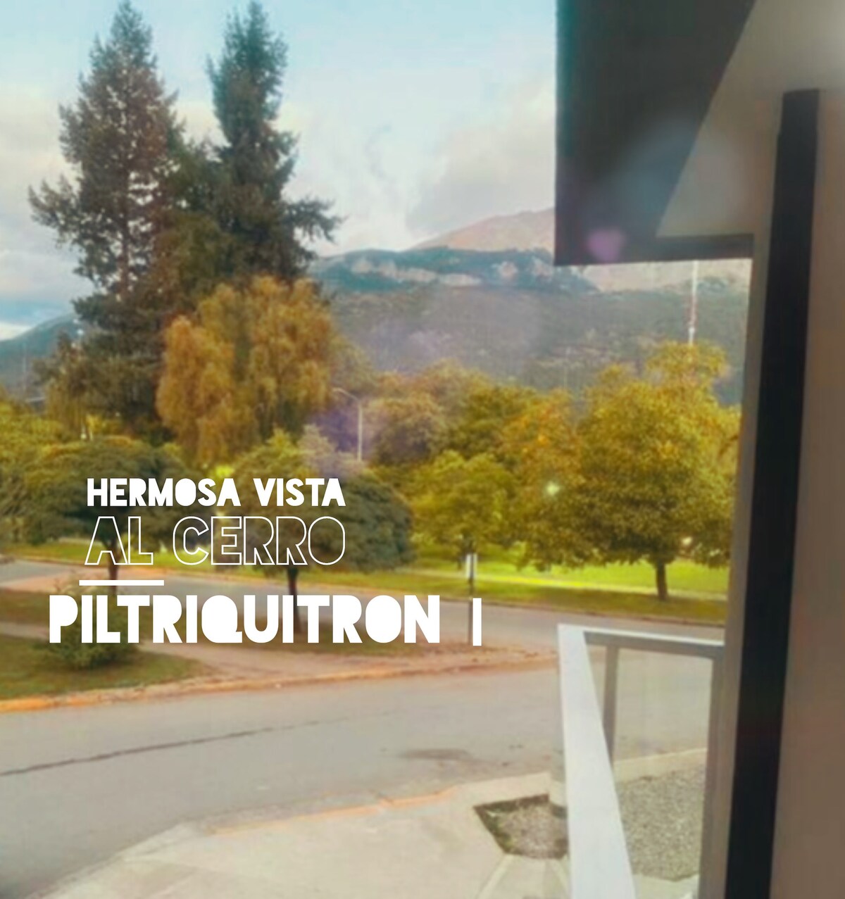 A view from the accommodation is presented, showcasing green trees and rolling hills leading to the Piltriquitrón mountain. The scene highlights a peaceful outdoor setting with a pathway visible, inviting exploration and engagement with the surrounding nature.