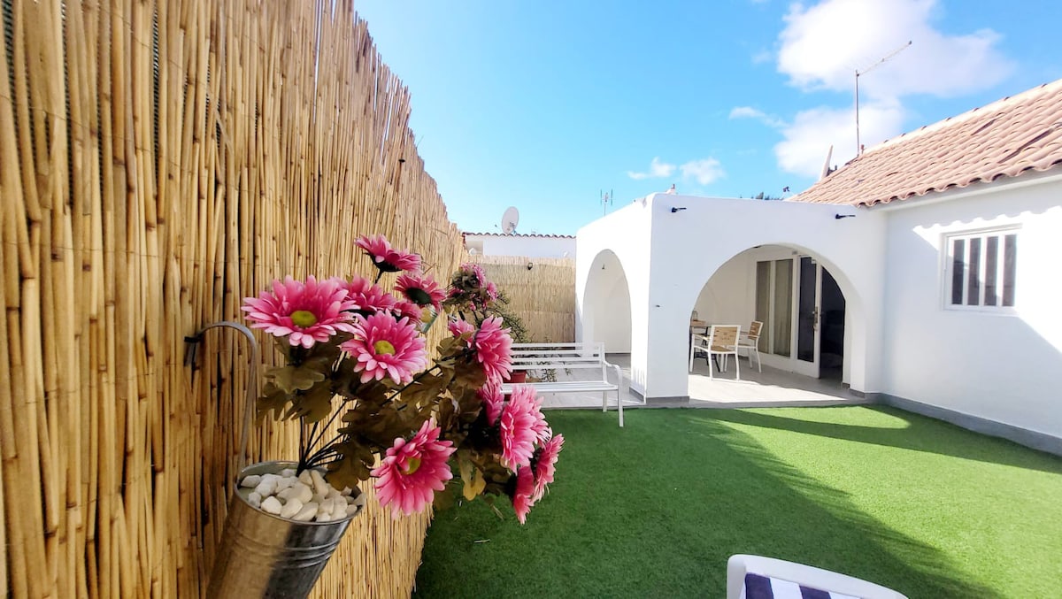 A private outdoor area features vibrant pink flowers in decorative pots, surrounded by natural bamboo fencing. A green grass lawn is complemented by white furnishings, including a seating area and a table, all set against a backdrop of a white-walled bungalow and blue sky.