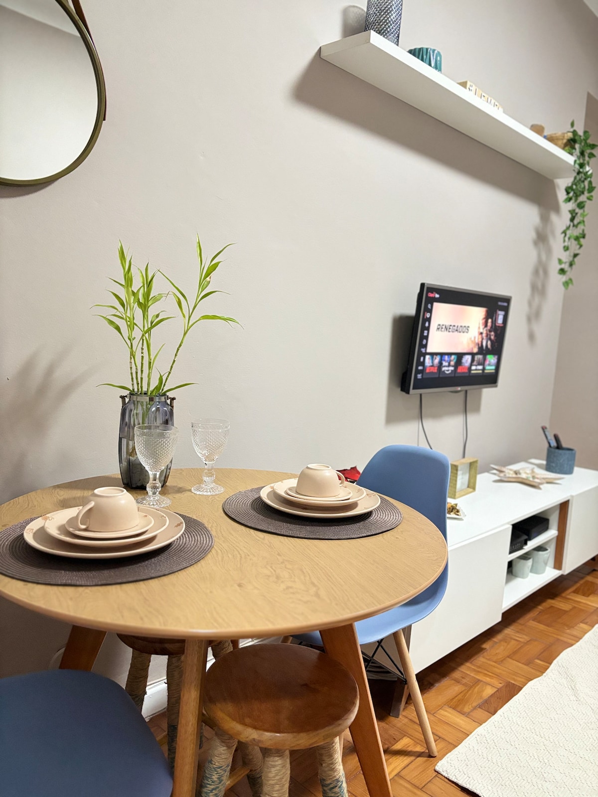 A dining area is highlighted by a round wooden table set for two. Neutral-colored plates and glasses are arranged atop woven placemats. A potted bamboo plant adds a touch of greenery beside a mounted TV on the wall. Light wooden shelving displays decorative items.