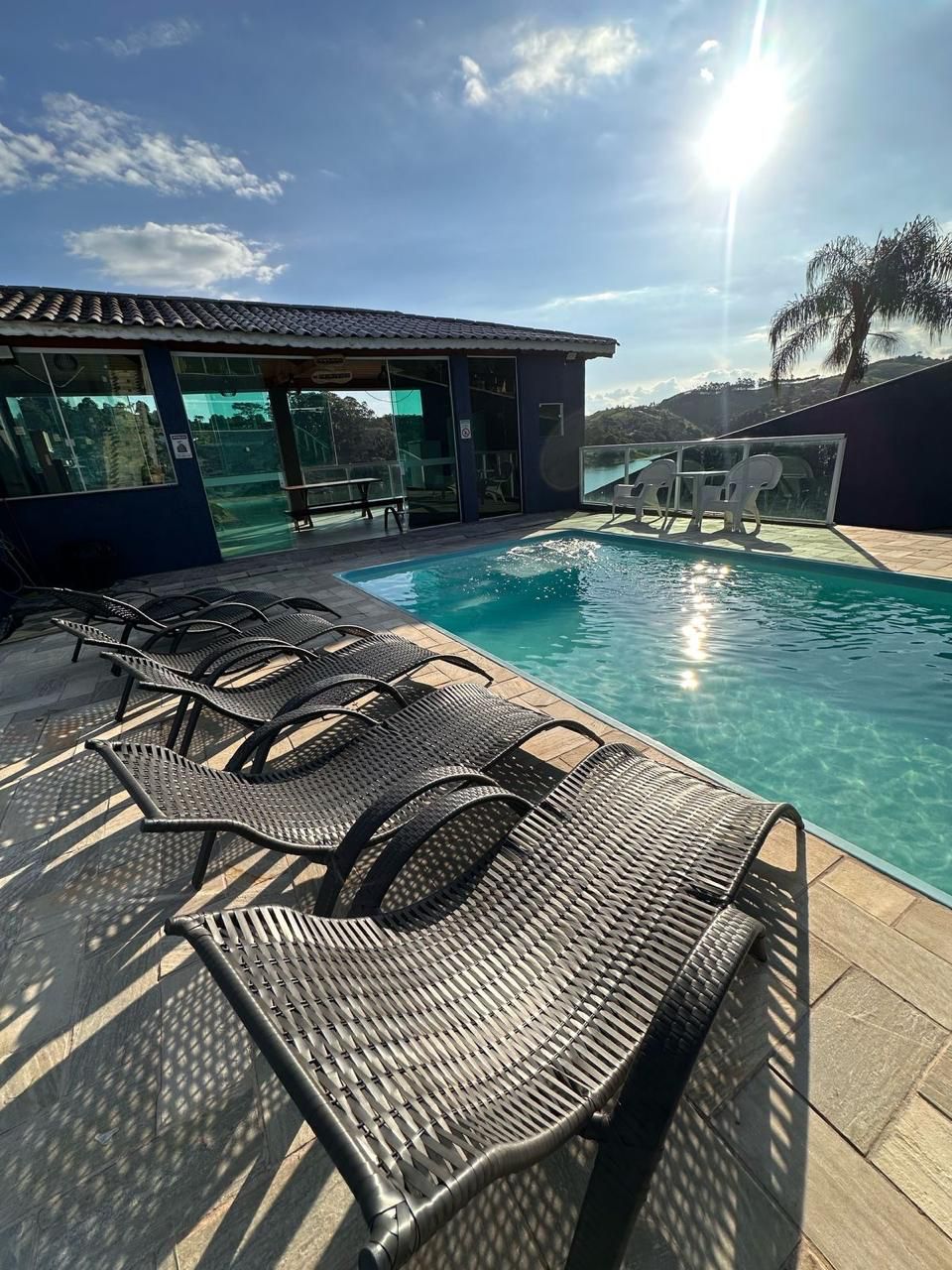 A large, crystal-clear swimming pool is surrounded by a spacious deck. Several lounge chairs are positioned nearby, offering a comfortable space for relaxation. The bright sunlight reflects off the water's surface, and a distant view of greenery is visible in the background.