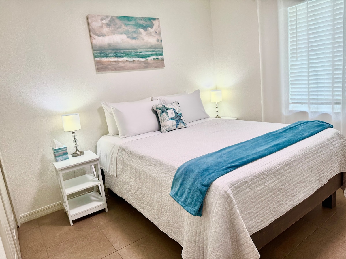 A serene sleeping area is highlighted by a spacious bed adorned with crisp white linens and a soft blue throw. Two bedside lamps provide gentle illumination, and a coastal-themed artwork hangs above the bed. Natural light filters through a window with white blinds.