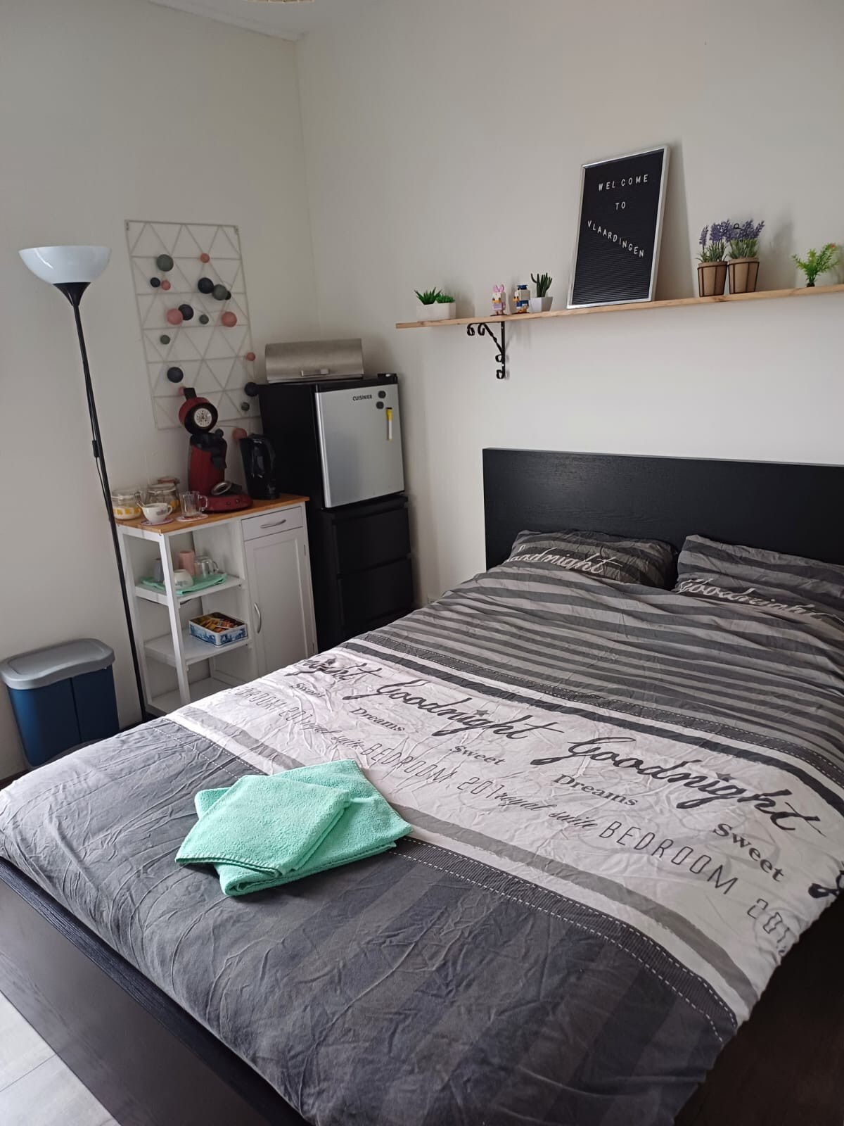 A well-organized bedroom features a spacious bed with a decorative duvet cover. Nearby, a mini-fridge and coffee station are available, along with a plant-lined shelf and soft lighting. A light green towel is neatly placed at the foot of the bed, adding a serene touch.