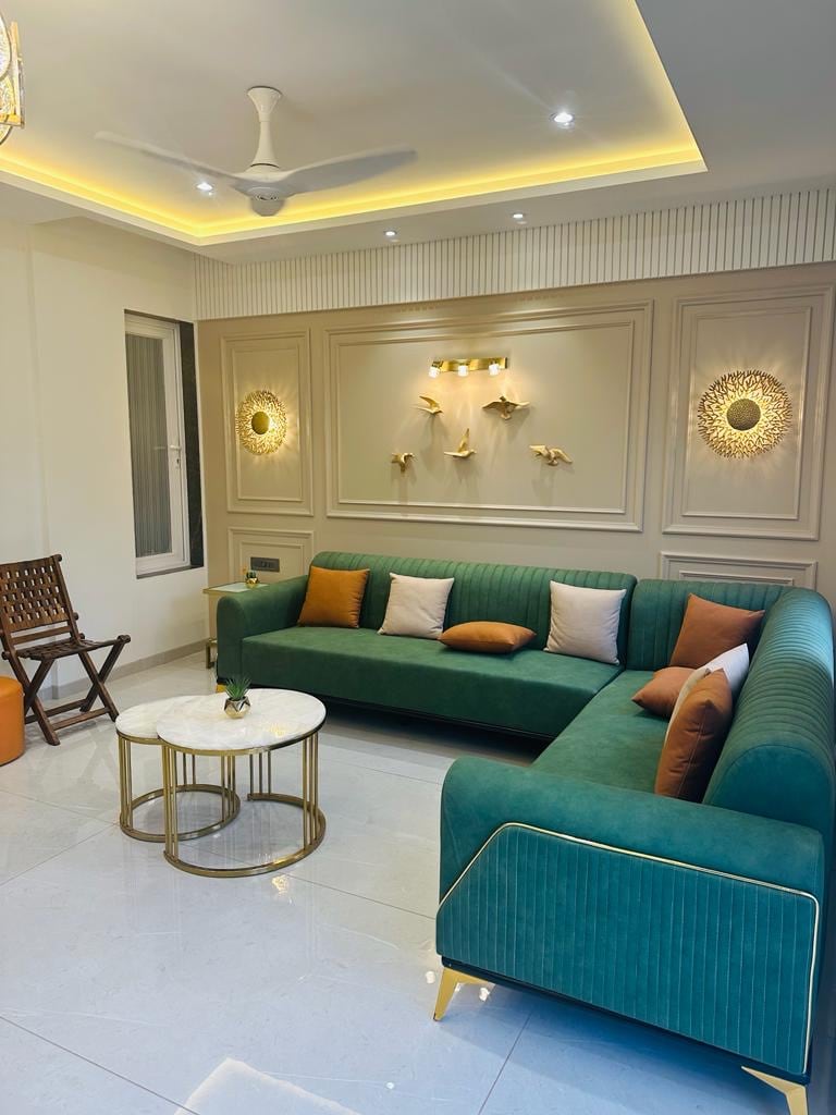 A modern living area is highlighted by a green sectional sofa adorned with neutral and warm-toned cushions. A round, white marble coffee table is positioned in front, complemented by decorative wall fixtures. Subtle lighting creates a calming ambiance, enhancing the room's contemporary design.