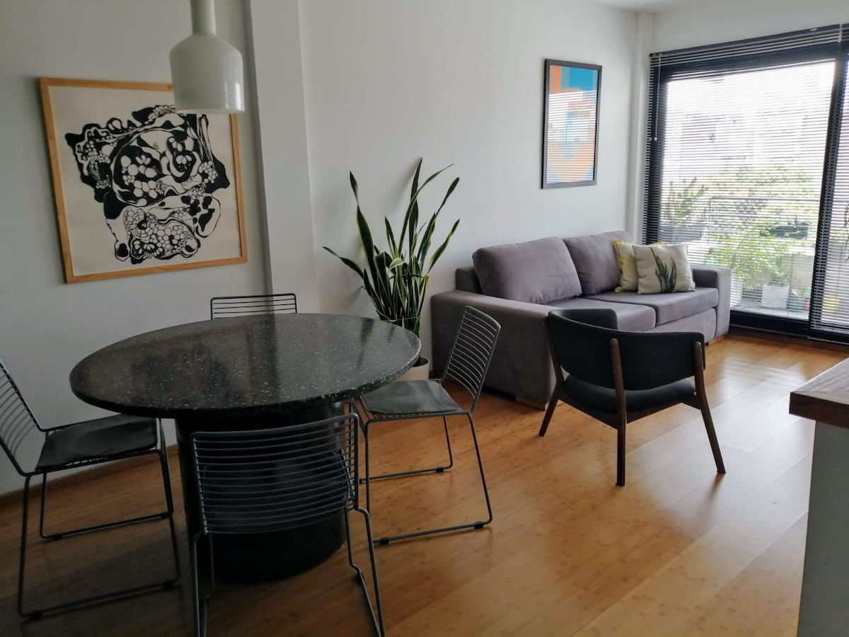 A dining area is presented with a round black marble table surrounded by six metal chairs. A cozy gray sofa with decorative pillows is visible beside large windows, which offer views of green plants outside. Artwork adorns the wall, adding a contemporary touch.