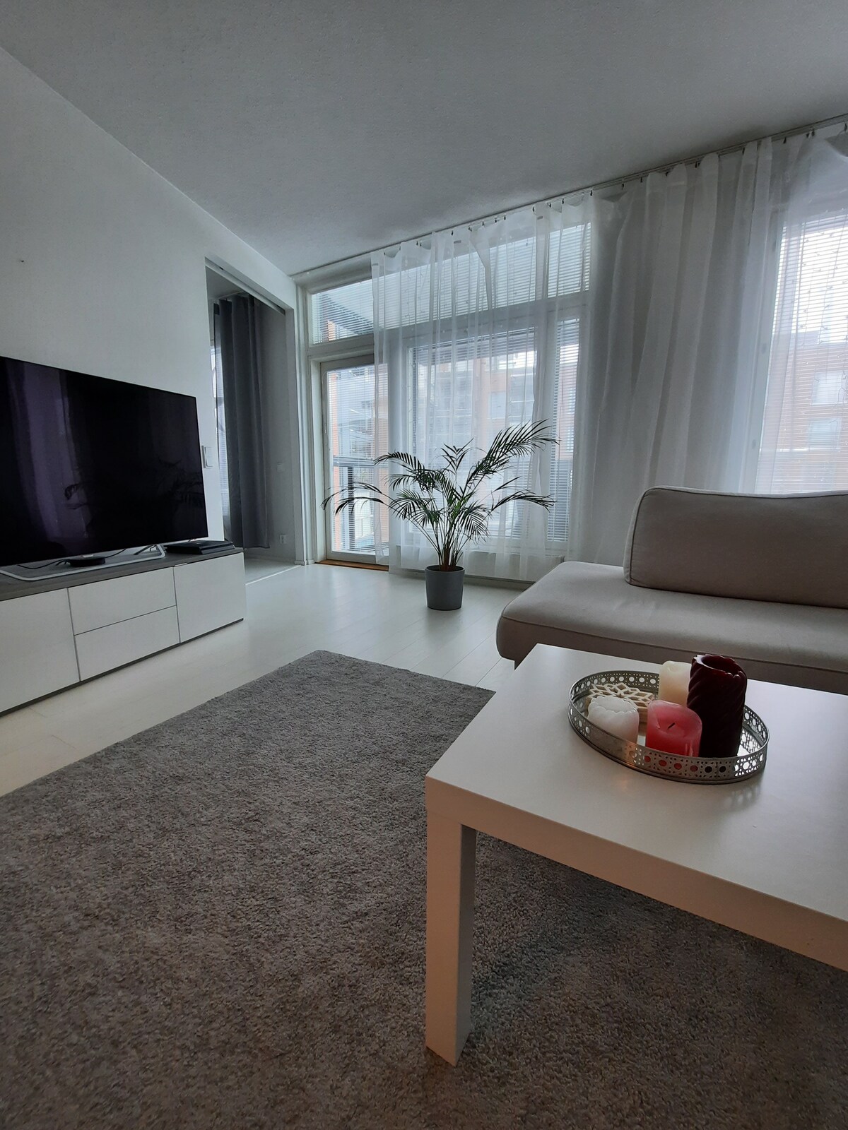 A bright living room features a spacious grey rug and a large light-colored sofa. A 70-inch smart TV stands on a white entertainment unit. Large windows are adorned with sheer curtains, allowing natural light to fill the room. A potted plant adds a touch of greenery.