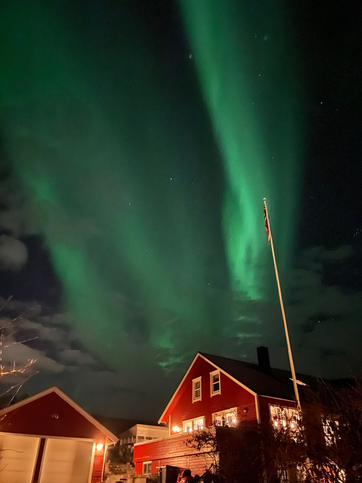 The night sky is illuminated by vibrant green northern lights, casting an ethereal glow above a red house. A flagpole stands in the foreground, with the house's warm lights visible through the dark, partly cloudy atmosphere.