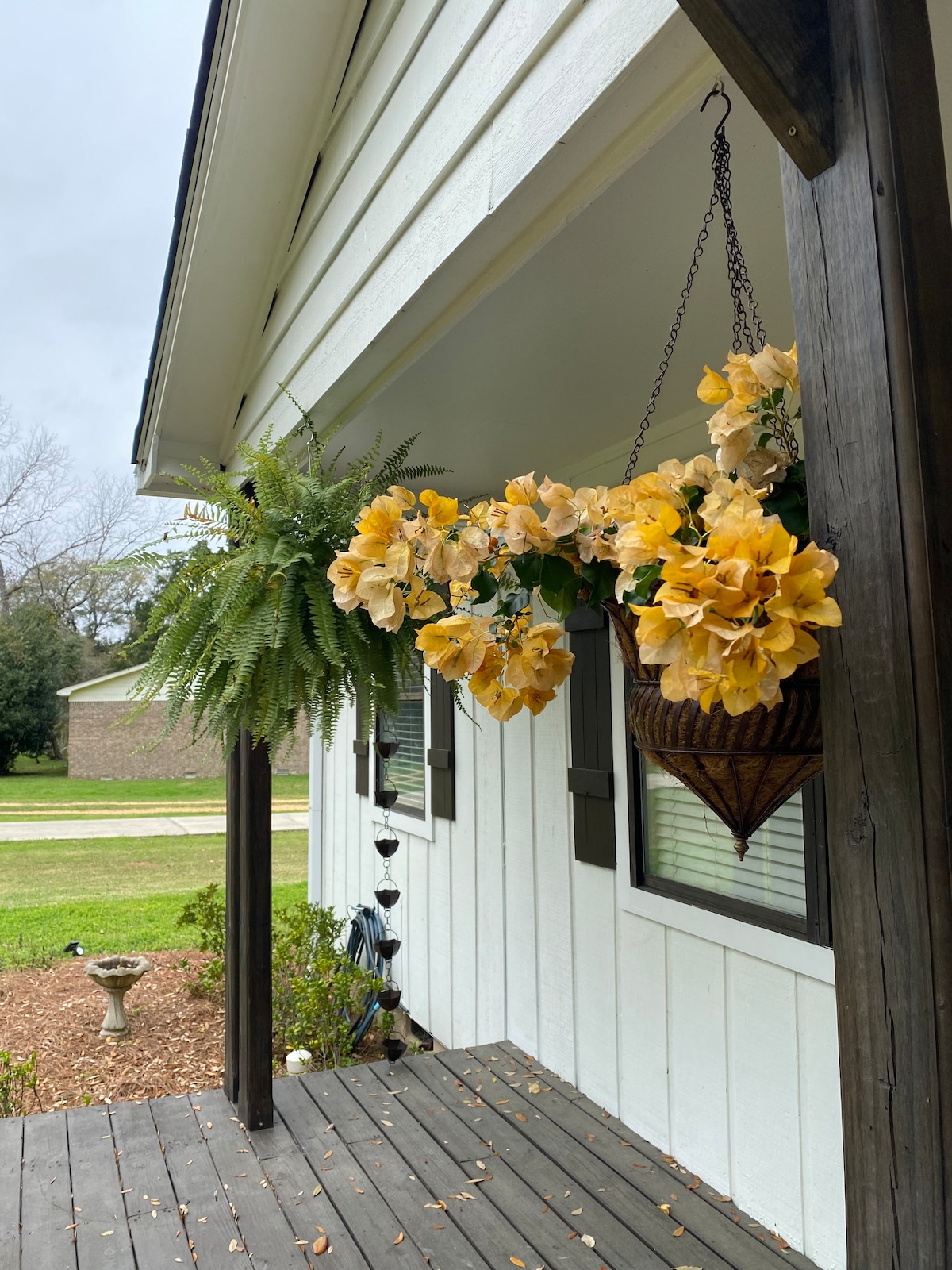 A welcoming porch area is adorned with a hanging basket filled with yellow flowers. Lush green ferns sway gently, complementing the natural beauty. The entrance features a wooden post and a clean, modern aesthetic, with windows allowing for ample natural light.