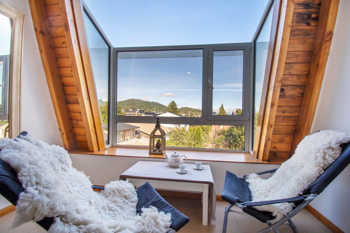 A cozy seating area is created by two comfortable chairs draped with soft throws, positioned near a large window. The expansive view highlights surrounding hills and trees, enhancing the bright, airy atmosphere. A small table holds tea cups, inviting relaxation and enjoyment of the scenery.