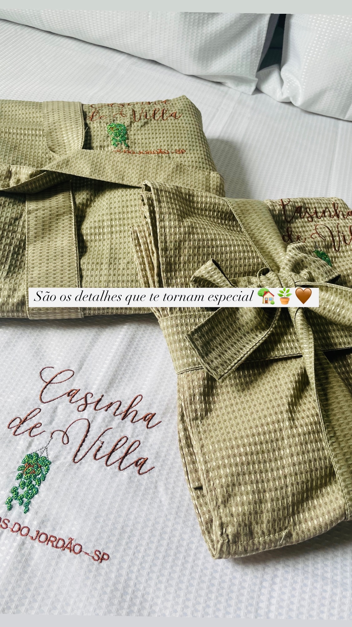 Two cozy robes are neatly arranged on a white bedspread featuring an embroidered design that reads 'Casinha de Villa.' The textured fabric of the robes adds a welcoming touch, enhancing the overall charm of the space.