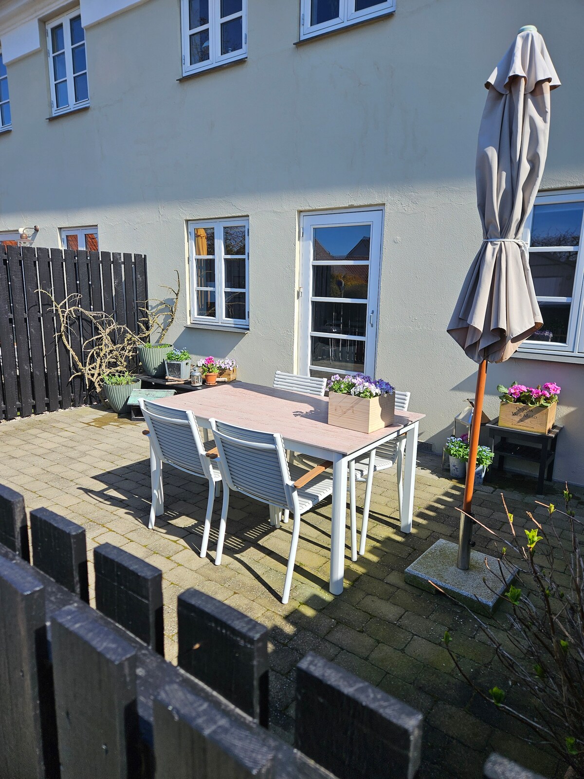 A private outdoor courtyard is presented with a wooden table surrounded by six white chairs. Potted plants with colorful flowers add life to the area. An umbrella is positioned next to the table, providing shade, with a black wooden fence enclosing the space.
