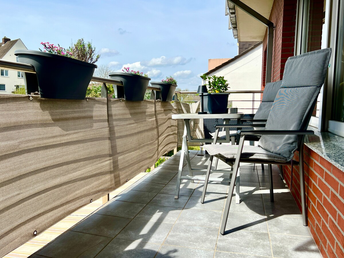 A balcony features two grey chairs and a table, providing a space for relaxation. Flower planters are positioned along the railing, adding a touch of greenery. The floor is tiled, enhancing the area for outdoor enjoyment.