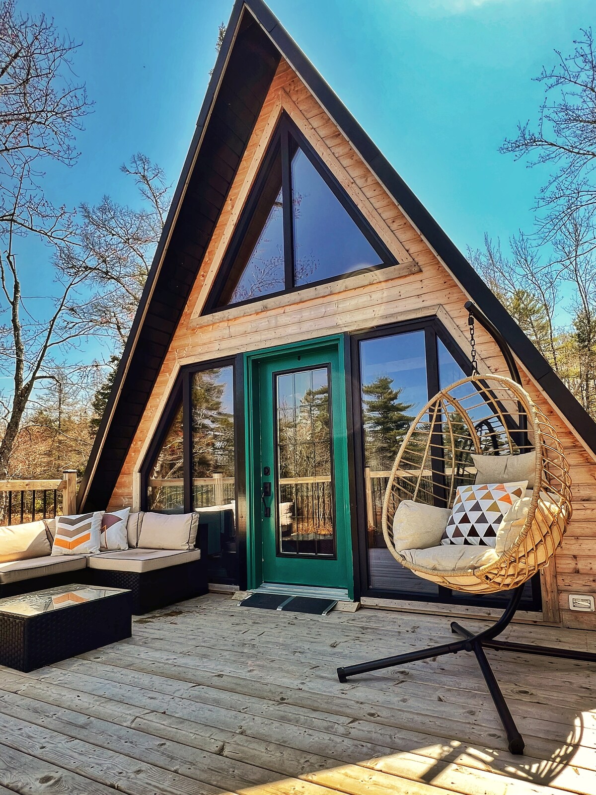The exterior of the A-Frame cabin features large triangular windows that allow natural light to fill the space. A cozy seating area is visible on the deck, complemented by a hanging egg chair, all set against a backdrop of trees and blue sky.