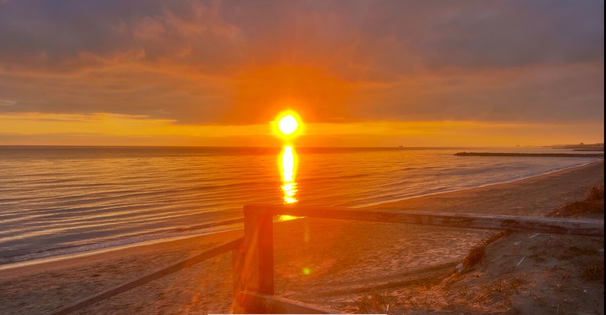 The serene coastline is depicted at sunset, with warm orange and yellow hues reflecting off the calm water. A sandy beach is visible in the foreground, framed by a wooden barrier leading towards the water's edge.