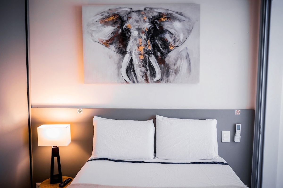 A cozy sleeping area features a spacious bed with crisp white linens and two large pillows. Above the bed, a striking elephant art piece adds a serene touch, while a bedside lamp provides gentle illumination.