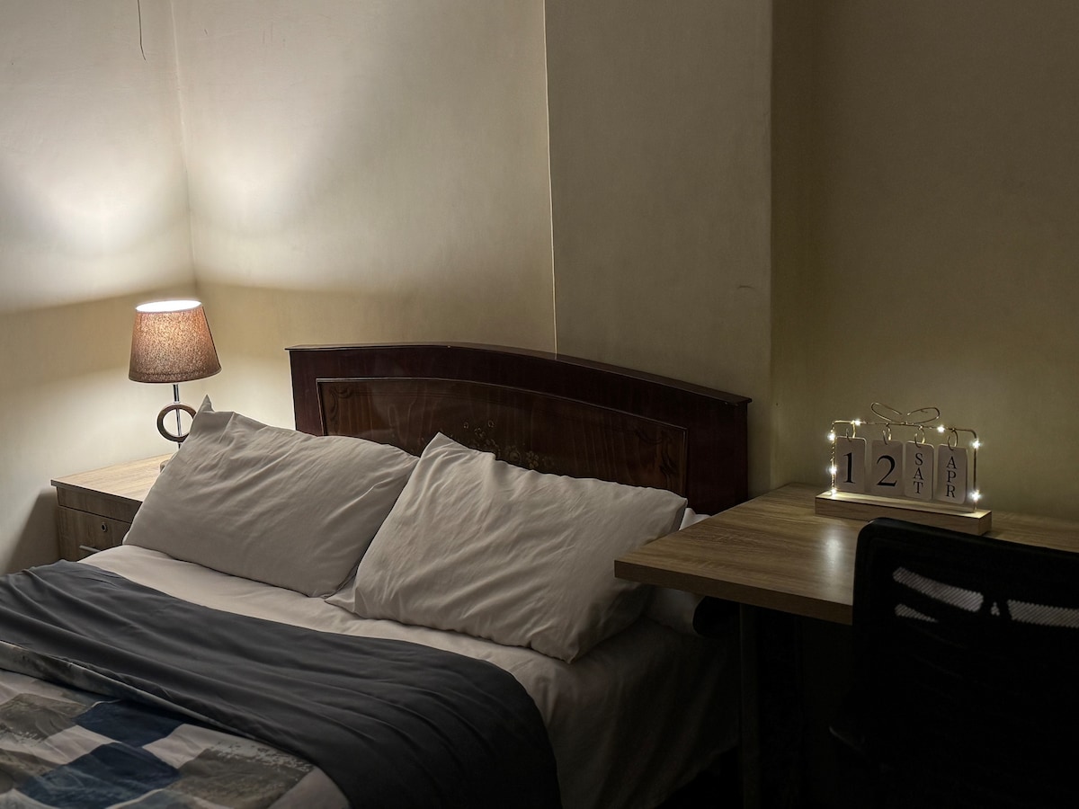 A cozy bedroom features a spacious bed with white pillows and a gray blanket. A simple wooden side table holds a warm lamp, casting a soft glow. A desk with a chair is positioned nearby, complemented by a decorative calendar.