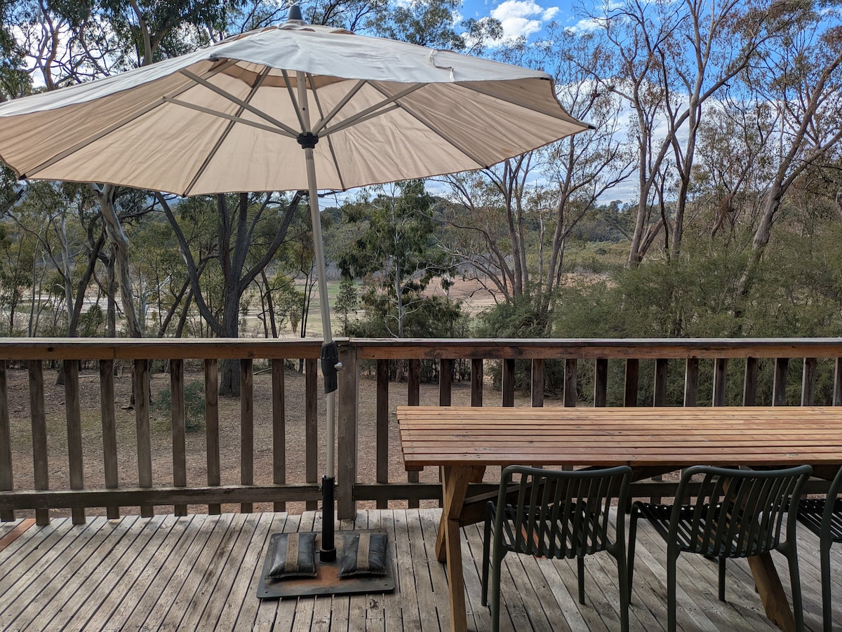 A spacious deck is shown with a large umbrella providing shade over a wooden table and four chairs. The surrounding bushland offers a scenic view, with trees visible in the background and a clear sky above.
