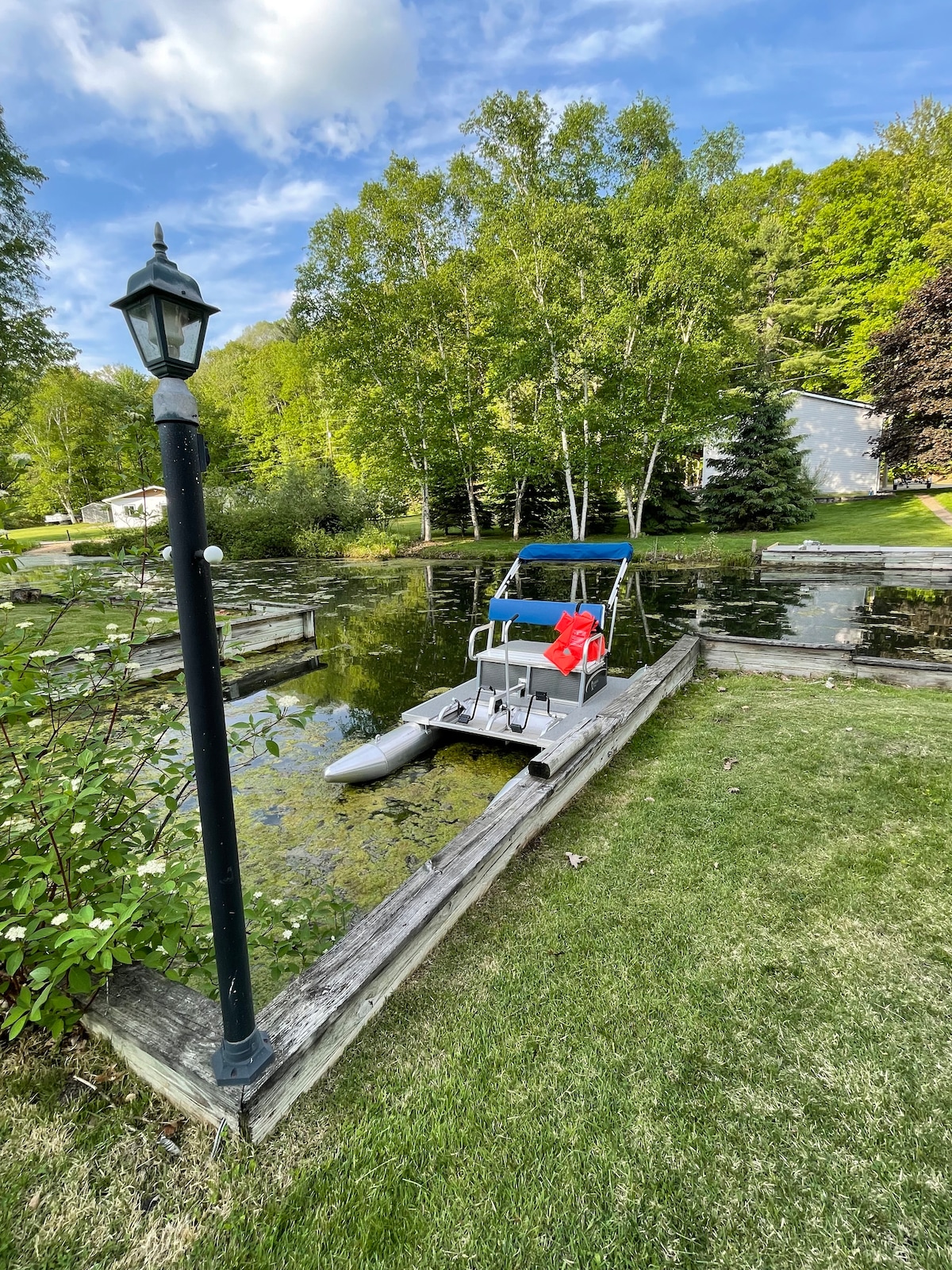 A small docking area is visible alongside a reflective inlet, featuring a pontoon boat secured at the end of wooden planks. Lush greenery surrounds the water, with light filtering through the trees. A lamp post stands near the edge, adding a subtle touch to the scenery.