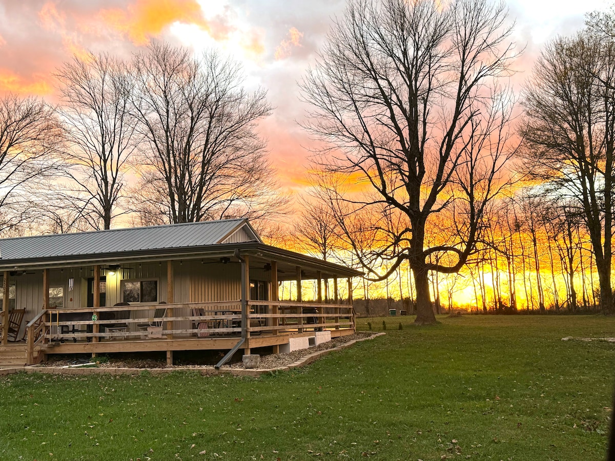 A farmhouse is set against a vibrant sunset, revealing colorful hues of orange and pink in the sky. The structure features a covered porch with wooden railings, surrounded by open grassy areas and tall trees, creating a serene rural landscape.