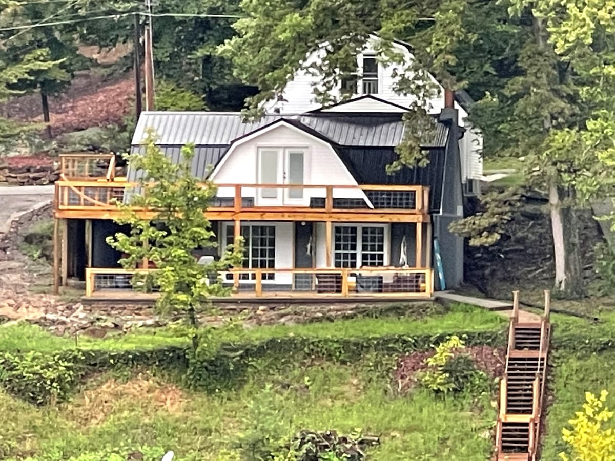 The unique three-level home is showcased from an elevated viewpoint, featuring a wrap-around deck and large glass doors that open to a welcoming outdoor space. Green surroundings and a staircase leading to the lower level highlight the tranquil lakeside setting.