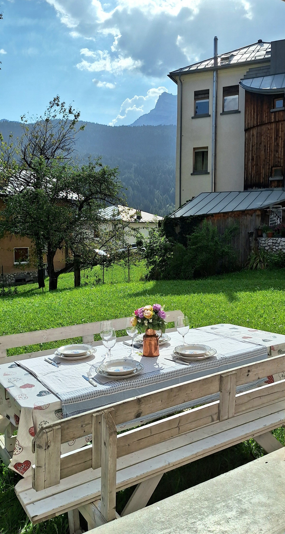 An outdoor dining area features a wooden table set with plates and glasses, surrounded by a grassy lawn. In the background, a multi-story building is visible along with trees and a mountainous landscape under a partly cloudy sky.